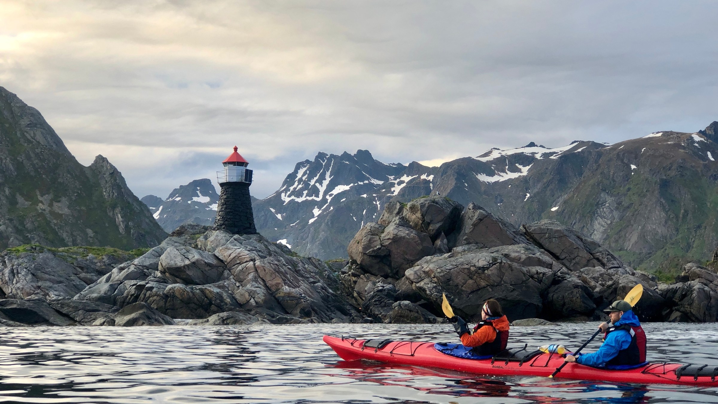 Two people kayaking near rocky shore with a lighthouse and mountains in the background.
