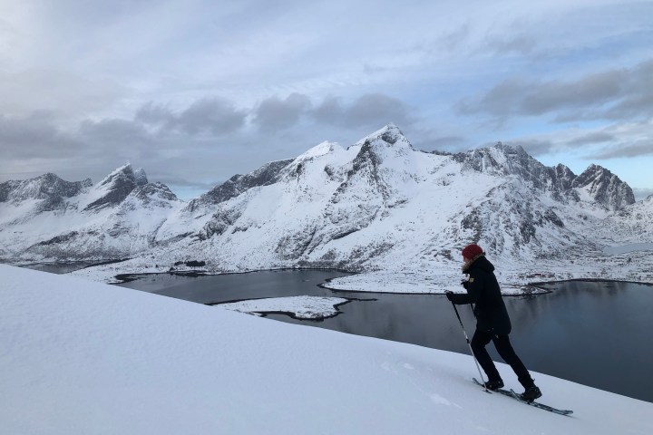 Person skiing uphill on snowy slope with mountains and lake in background.