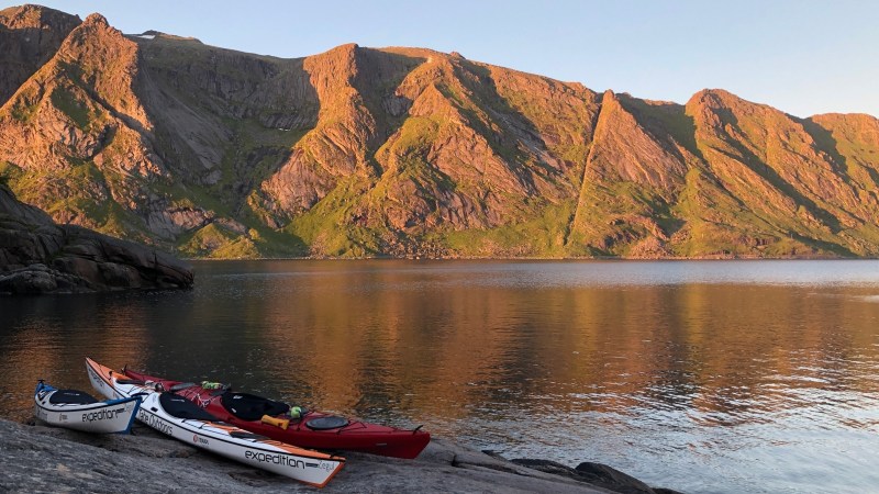 Kayaks on rocky shore with sunlight on mountain and calm water reflecting the scene.