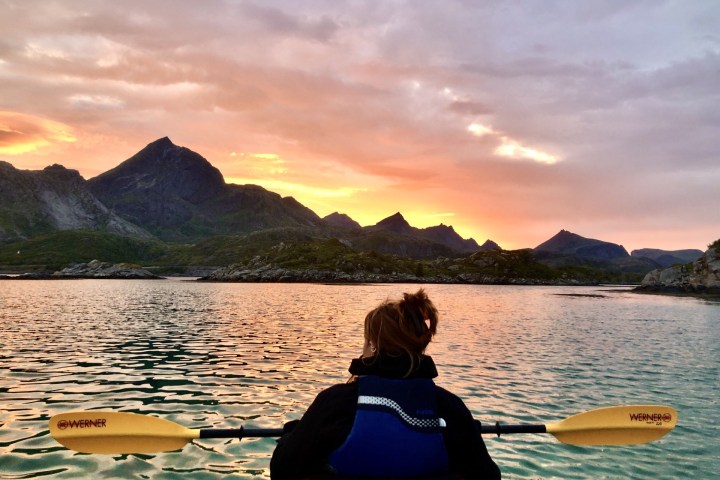 Person kayaking on a lake at sunset with mountain views.