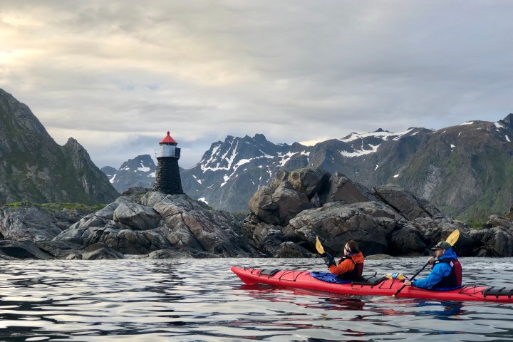 Two kayakers paddle near a rocky island with a lighthouse against a mountainous backdrop.