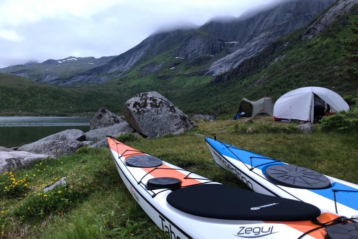 Two kayaks near tents on grassy shore with mountains and mist in the background.