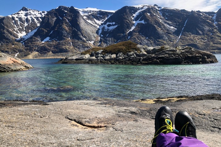 Person's feet overlooking a rocky mountain landscape and clear lake under a blue sky.
