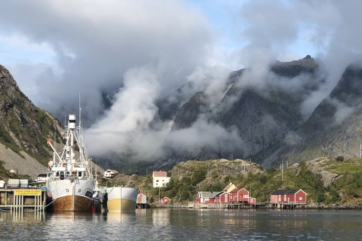Boats docked by red houses with mountains and clouds in the background.