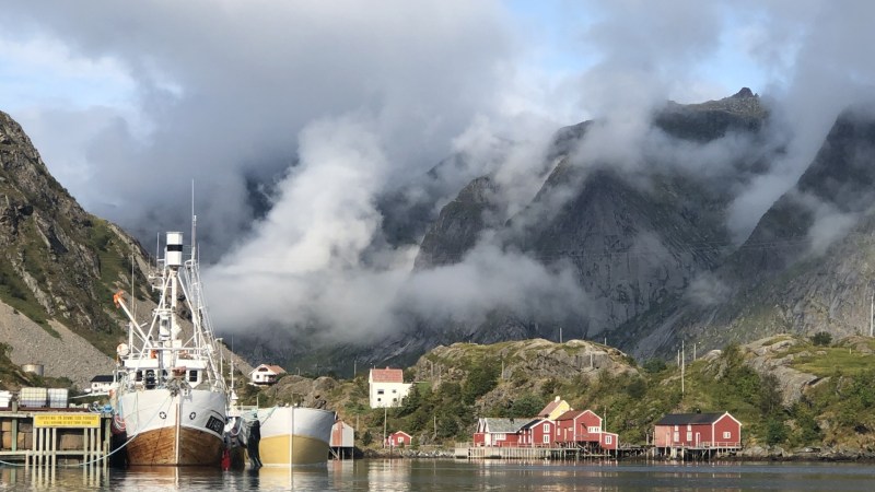 Boats docked by red houses with mountains and clouds in the background.