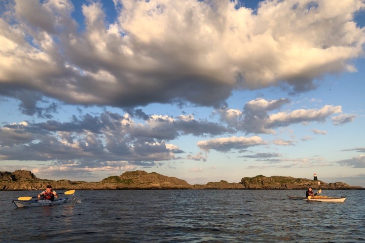Two kayakers paddle on calm water under a blue sky with large clouds and distant rocky islands.