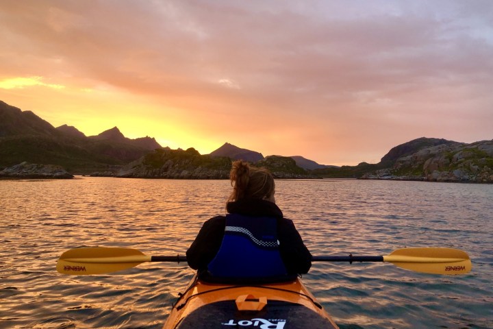 Person kayaking on a lake with a sunset and mountains in the background.