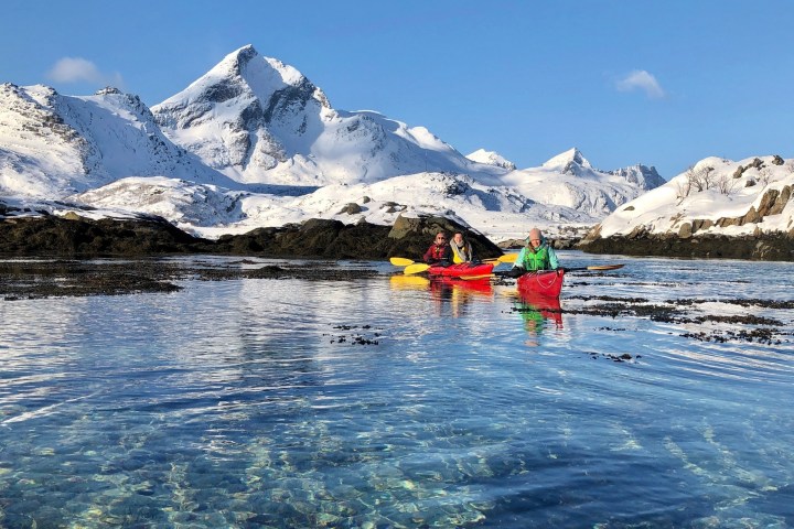 Three kayakers paddle in clear water with snowy mountains in the background.