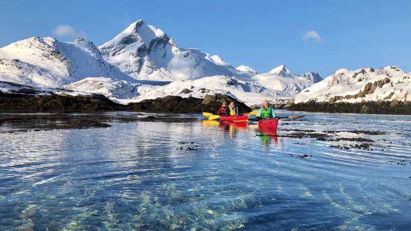 Three kayakers paddle in clear water with snowy mountains in the background.