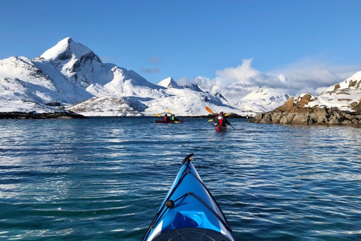 Kayakers in a blue ocean surrounded by snowy mountains under a clear blue sky.