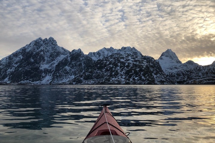 Kayak on calm water facing snowy mountains under cloudy sky at sunrise.