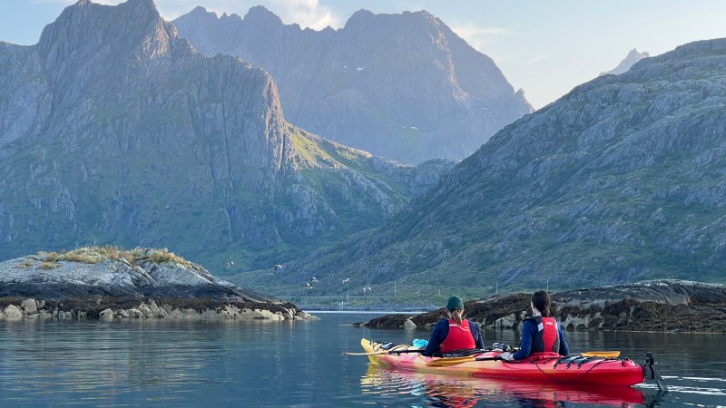 Two people in a red kayak on a calm lake, surrounded by mountains.