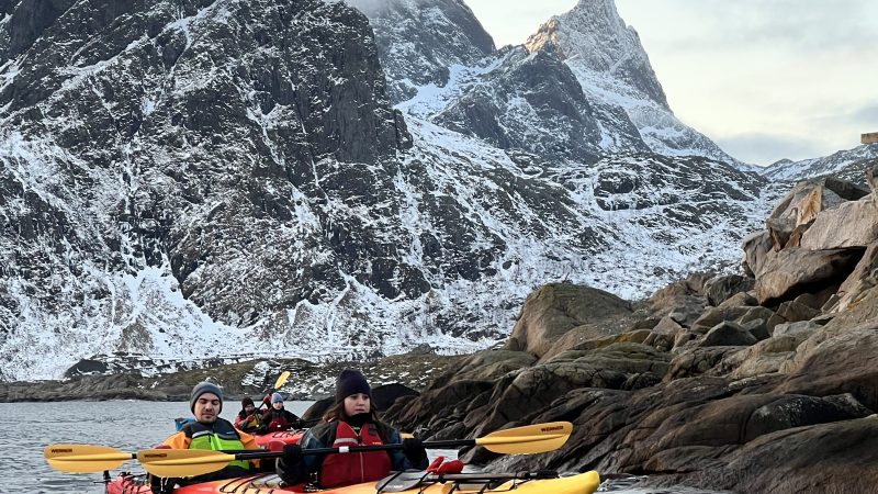 Kayakers in colorful kayaks on water with snow-capped mountains and rocky shoreline in the background.
