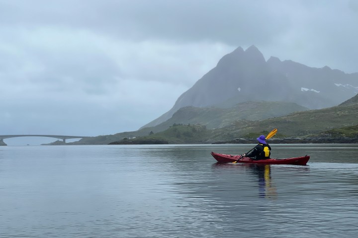 Person kayaking on calm water near a bridge and mountains on a cloudy day.