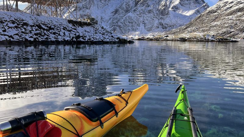 Two kayaks on a clear lake with snow-covered mountains and blue sky in the background.