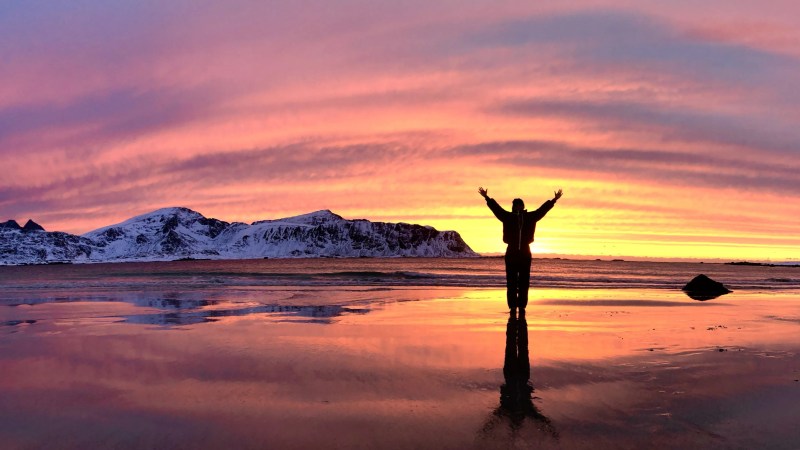 Silhouette of person with arms raised on a beach at sunset, reflecting on wet sand.