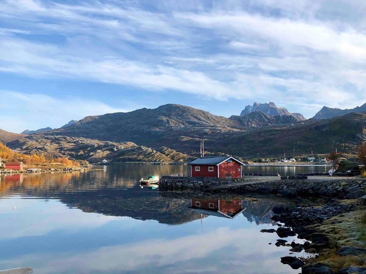 Red cabin by a calm lake reflecting mountains and a cloudy sky.