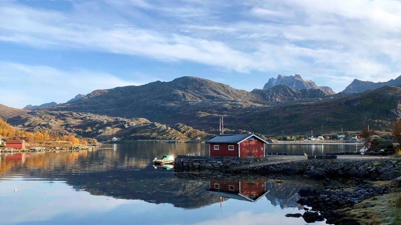 Red cabin by a calm lake reflecting mountains and a cloudy sky.