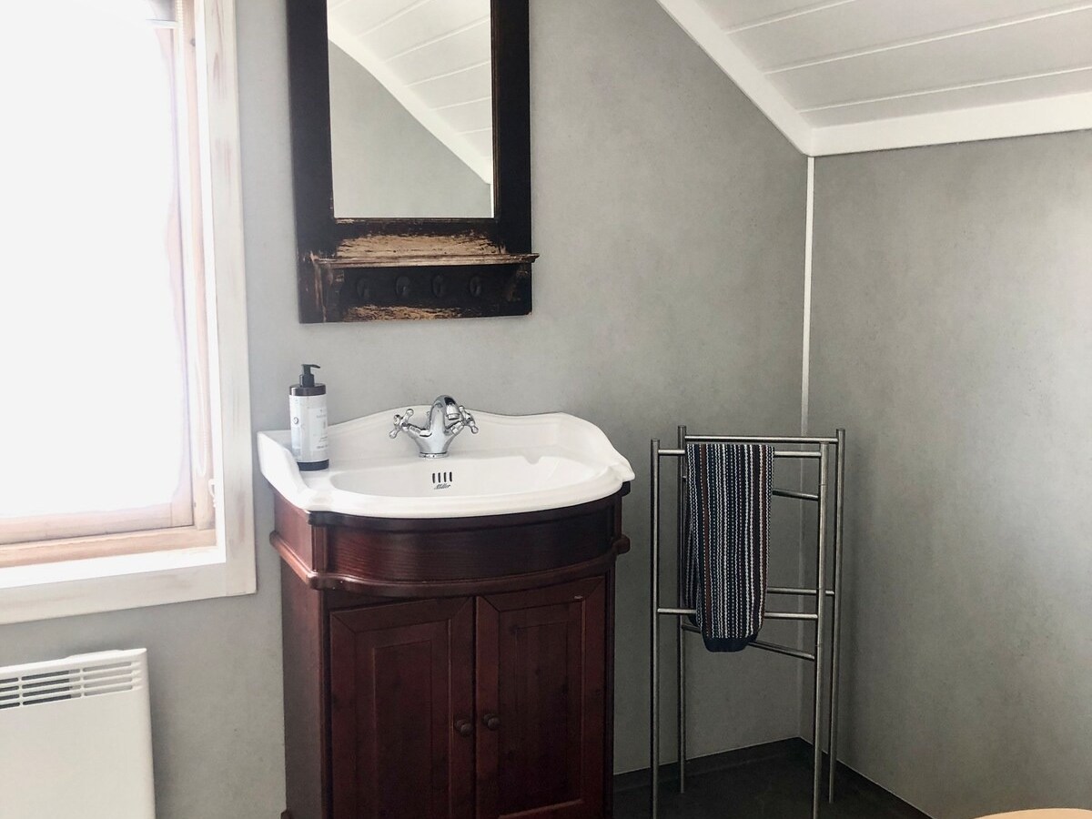 Bathroom with sink, mirror, towel rack, and wooden toilet lid under slanted ceiling.