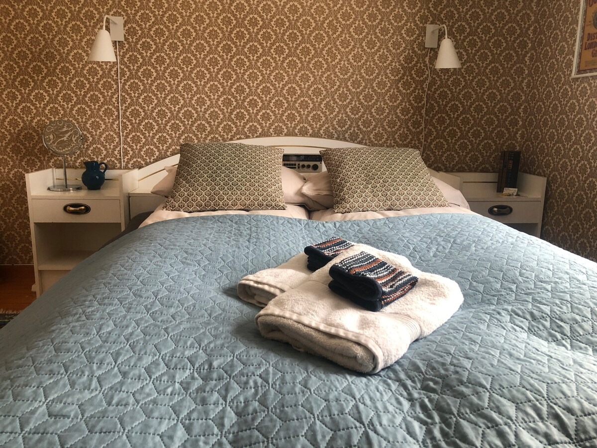Cozy bedroom with blue quilt, patterned pillows and folded towels on bed, vintage wallpaper in background.