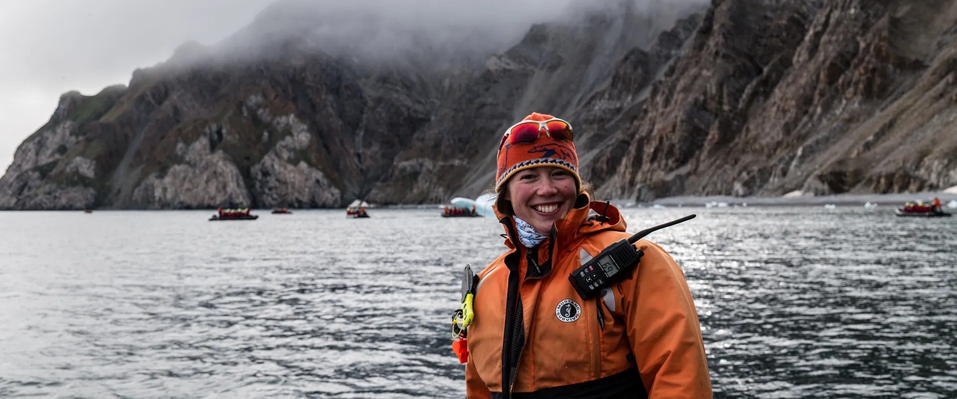 Smiling person in orange jacket on boat with rocky, foggy cliffs in background.