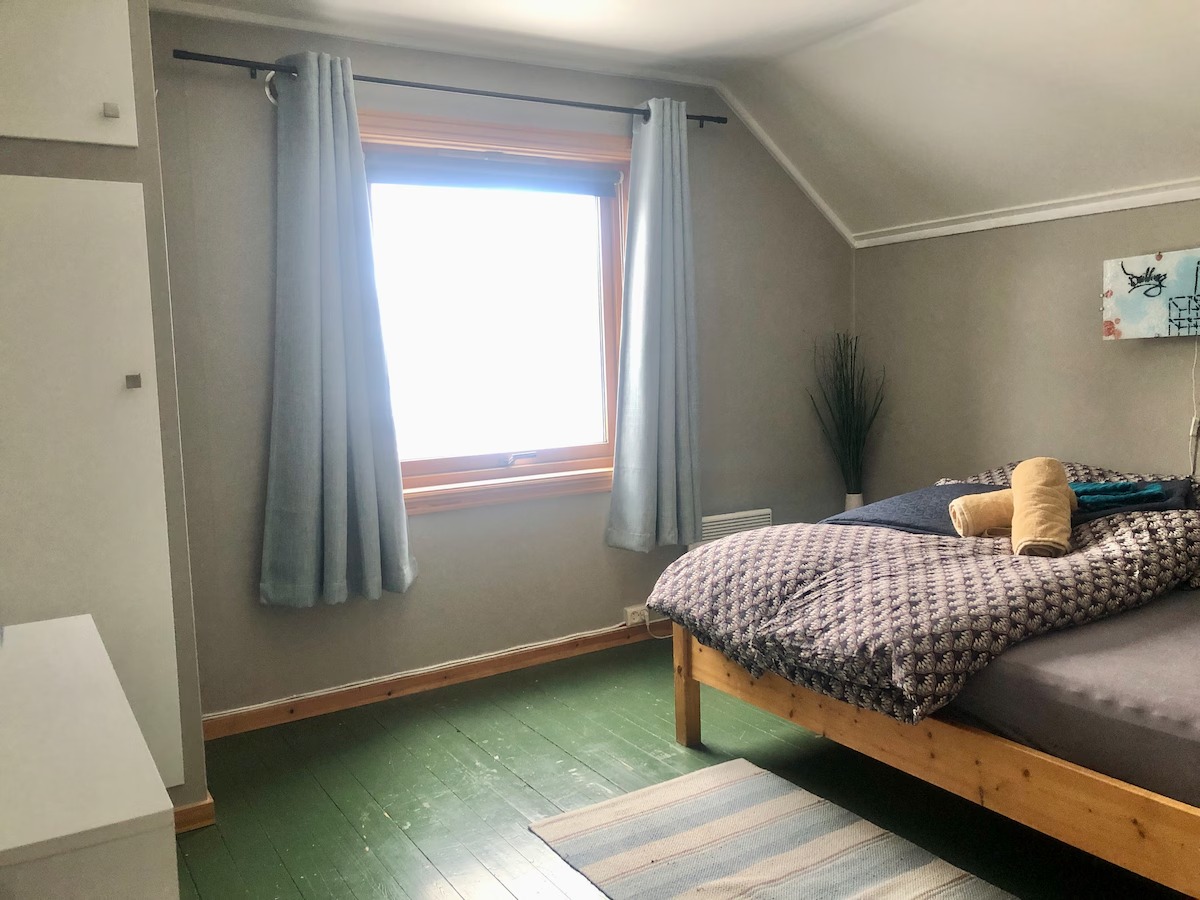Bright bedroom with wooden bed, blue curtains, and green floor, featuring a large window.
