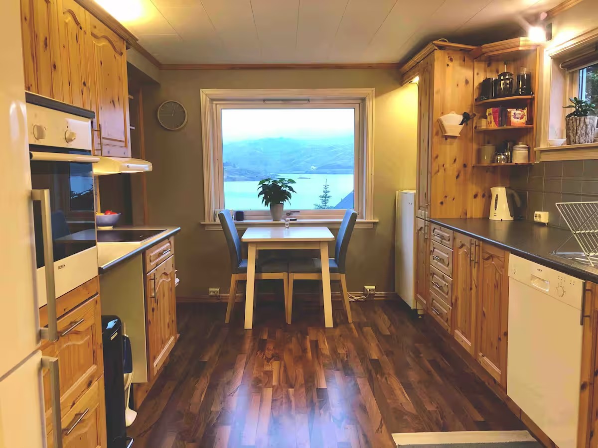 Cozy wooden kitchen with dining table by window, overlooking scenic lake and mountains.
