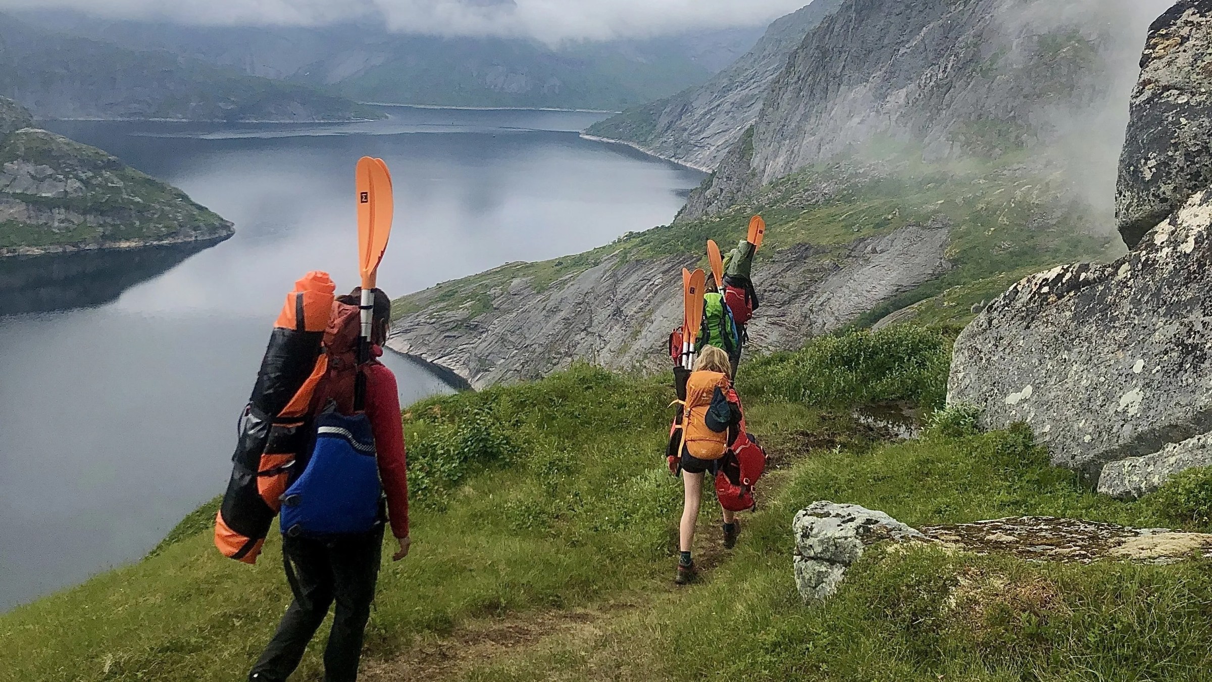 Hikers with backpacks and paddles walking along a lake path in mountainous terrain.