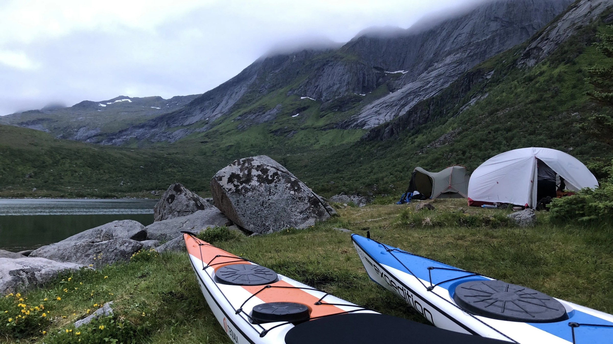 Two kayaks by tents in a rugged mountain landscape and cloudy sky.
