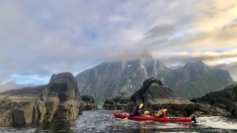 Two people kayaking between rocks with mountains in the background under a cloudy sky.