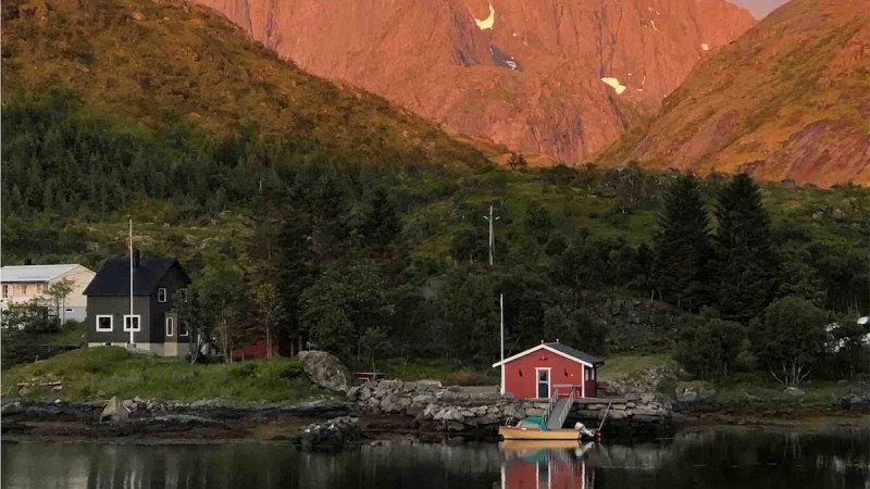 Mountain backdrop at sunset with red cabin and calm lake reflection.